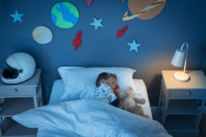 Child sleeping with teddy bear in space-themed bedroom, promoting healthy sleep for mental health and learning.