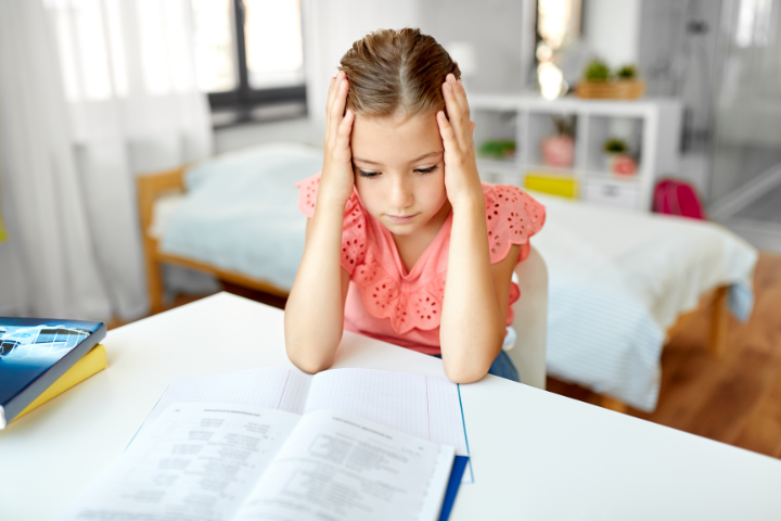 A young girl sits at a desk with her head in her hands, looking worried while studying — illustrating childhood anxiety and stress.