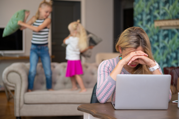 A tired mother sits at a table with her laptop, head in hands, while two children play noisily behind her on the sofa — a scene reflecting the need for stress management for parents.