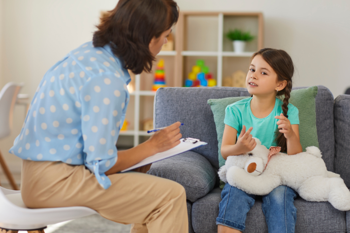 A young girl sits on a couch holding a white stuffed toy while talking to a woman with a clipboard, likely a therapist or counselor who shares support.