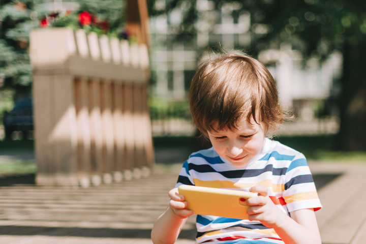 Child sitting outdoors on a sunny day, focused on a yellow tablet, illustrating summer screen-time balance.